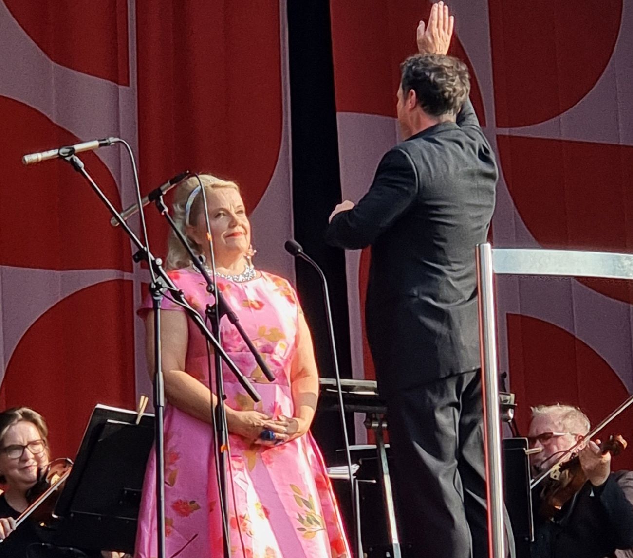 Tampere Filharmonian Puistokonsertti Tampereen Sorsapuistossa 9.8.2025. Tampere Filharmonian Puistokonsertti on yksi varmimmista loppukesän merkeistä Tampereella. Perinne jatkuu vähintäänkin entisellä voimallaan, vaikka ylikapellimestari vaihtui takinsaumansa taannoin halkaisseesta Santtu-Matias Rouvalista brittiläiseen Matthew Hallsiin. Kesäisen kepeä ohjelmisto saa kenen tahansa kuulijan tuntemaan itsensä melkein klassisen musiikin asiantuntijaksi. Tällä kertaa esimerkiksi konsertin alkupään Edvard Griegin Peer Gynt -sävellykset Aamutunnelma ja Vuorenkuninkaan luolassa ja päätteeksi soinut Maurice Ravelin Boléro kuuluvat kaikkein tunnetuimpiin klassisiin kappaleisiin. Olisiko ohjelma tänä vuonna rakennettu jopa aiempaa vieläkin helpommin kuunneltavaksi Pjotr Tshaikovskin Prinsessa Ruususineen ja Pähkinänsärkijöineen sekä Sergei Prokofjevin Romeo ja Julia -sävelmineen? No, mikäs siinä, ei kai se haittaa, jos yleisö tykkää ja saa hyvän mielen. * * Sorsapuiston nurmikoille oli kertynyt noin 10 000 kuulijaa. Sää oli ehkä paras mahdollinen: ei liian kuuma, ei liian kylmä, aurinko paistoi kirkkaalta taivaalta, eikä sateesta tai edes pikkuruisesta ripauksesta ollut tietoakaan. Myrskyn nostivat ainoastaan Jean Sibeliuksen konserttisarjan osat. Muistissa on vielä takavuosien ankean sateinen ja kalsea konserttipäivä, jota ei minkäänlainen soitanta saanut lämpenemään. Lienevätkö edes muusikoiden sormet välttyneet kohmettumiselta. Nyt tunnelma sykki leppoisana, lämpöisenä ja nautittavana. Tampere Filharmonia on huippuvedossa, ei epäilystäkään. Se on valinnut 95-vuotisjuhliensa teemaksi rakkauden ja suuret tunteet. Eräs ulkomaalainen konserttiin osunut vieras hämmästyi äimäksi asti ”maakuntaorkesterin” huipputasosta ja aikoi ryhtyä selvittämään halpojen lentolippujen ostamista päästäkseen mahdollisimman useasti Tampere-talon saliin. KUVA Sopraano Anu Komsi lauloi Tampere Filharmonian solistina. Kuva: Aila-Liisa Laurila Nyt Puistokonsertin solistiksi kutsuttu sopraano Anu Komsi esiintyi kahteen otteeseen. Aluksi kuultiin Oskar Merikannon Päivyt paistaos ja Antonín Dvorákin Rusalka-oopperan laulu kuulle. Loppupuolella hän tunnelmoi Erkki Melartinin Minä metsän polkuja kuljen -kappaleen ja Erna Tauron iki-ihanan Syyslaulun tahtiin. Rusalkan laulussa Komsi pääsi antamaan näytteen oopperadiivan huikeimmista aariataidoista. Ääni lienee kantanut rautatien toiselle puolelle asti. Sorsapuiston lehvistö raikui. Mukavasti Komsi kertoi myös harvoin esitetyn Rusalka-oopperan taustoista, kuten sen, että tapahtumat on sijoitettu Viipuriin. * * Yksi loistopuoli Filharmonian Puistokonsertissa on, että se tutustuttaa klassisen musiikin saloja myös lapsiyleisölle. Jo esitysaika kello 18–20 sopii mainiosti pikkuväen päivärytmiin. Paikoillaan he eivät jaksa koko aikaa oikein istua, mutta tanssivat ja juoksentelevat tenavat näyttävät herättävän enemmän hymyjä kuin närkästystä. Samaan aikaan Tampere-talossa järjestetty Muumi 80 – Suuri syntymäpäiväjuhla toi kaikki Muumien ja Tove Janssonin ystävät paikalle. Runsas ilmaisohjelma ja lapsille suunnatut aktiviteetit takasivat tungoksen taloon. Osa jatkoi päivää konsertissa. Aila-Liisa Laurila KUVA Dj Juissi on Filharmonian vakiolämppäri. Kuva: Aila-Liisa Laurila